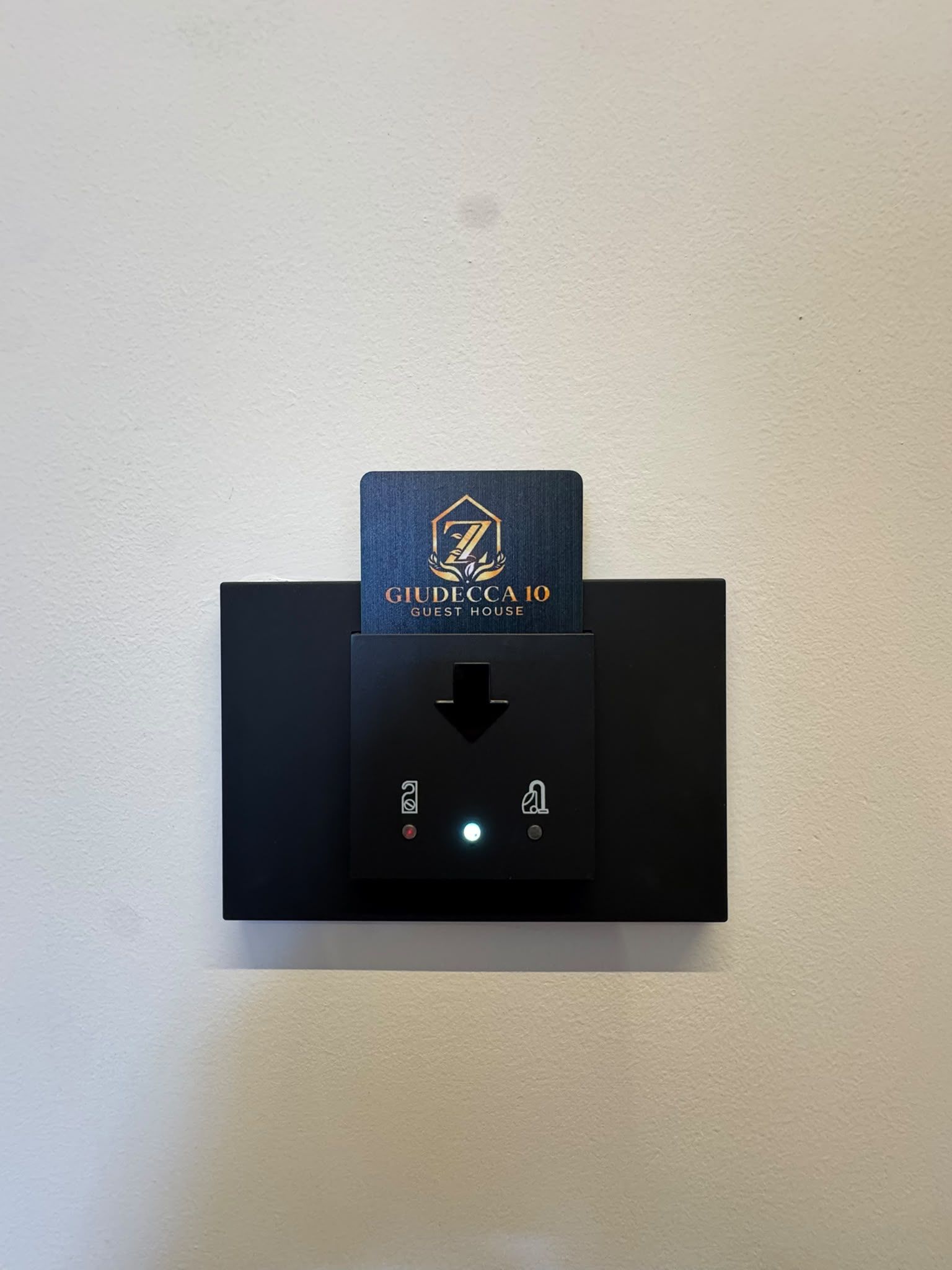 Eine wandmontierte Bedienkonsole mit Kartenhalter, der 'GIUDECCA 10 GUEST HOUSE' zeigt, und einer digitalen Schnittstelle mit Pfeilen und einer Leuchtanzeige.