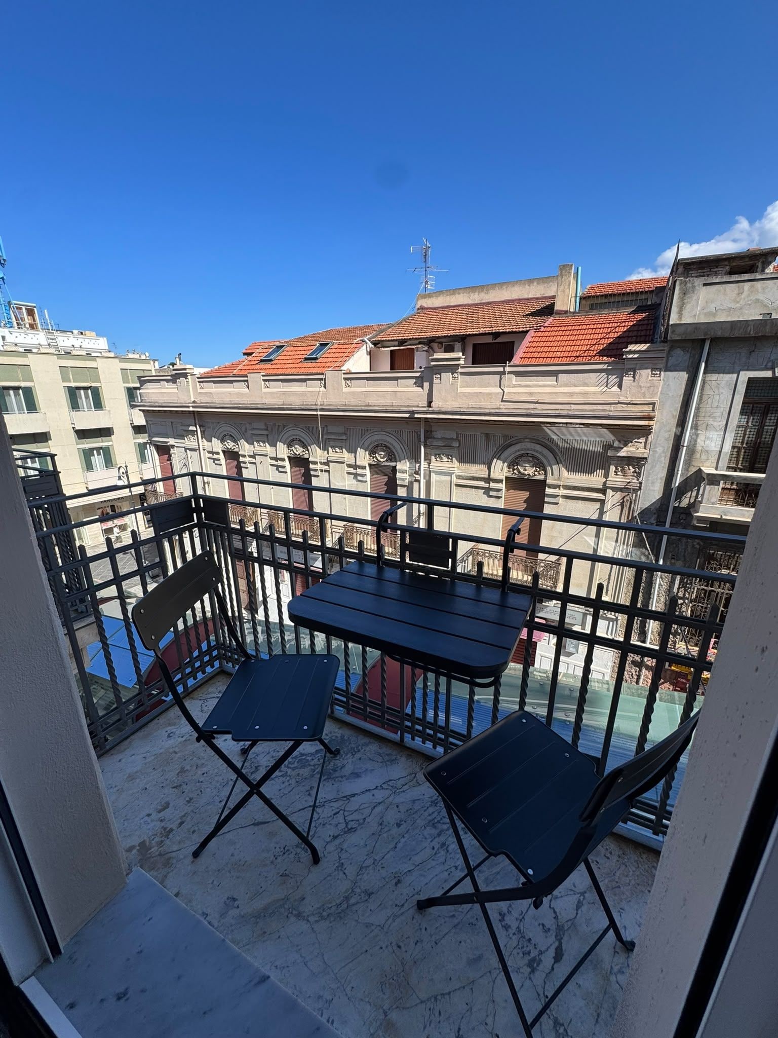 Ein Balkon mit Tisch und zwei Stühlen, mit Blick auf eine Stadtlandschaft mit roten Ziegeldächern.