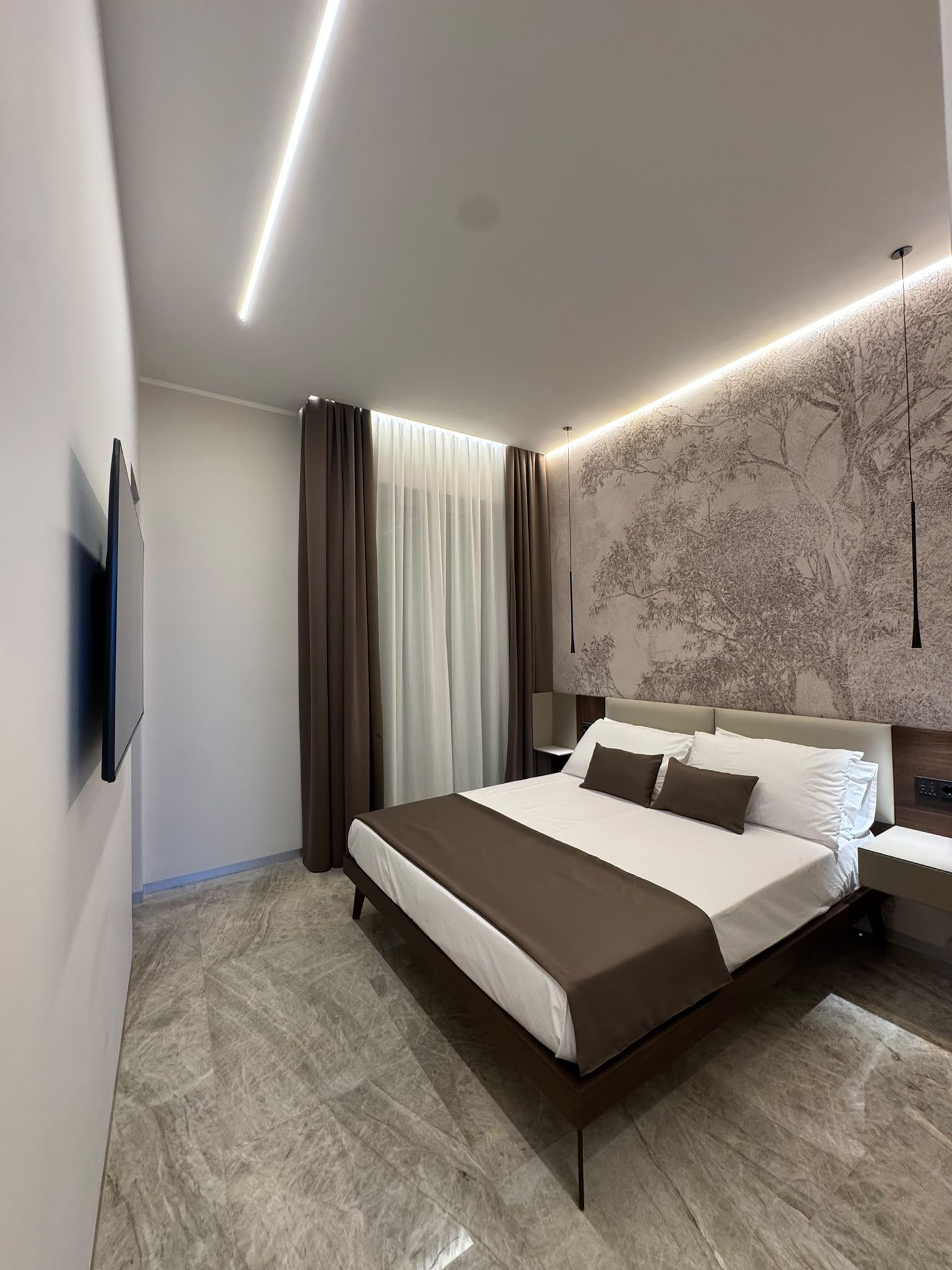 Une chambre moderne avec un grand lit, un téléviseur à écran plat fixé au mur et une fresque murale représentant un arbre à des fins décoratives.