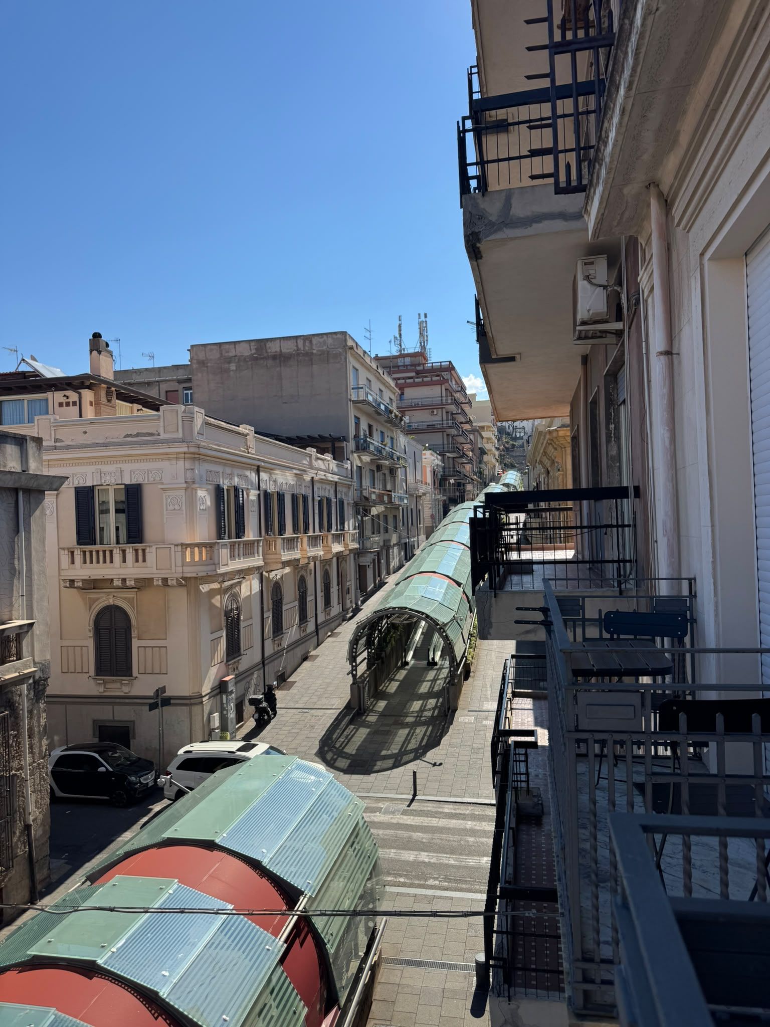 A narrow street with buildings on both sides, a parked car, and a green vehicle with a curved roof.