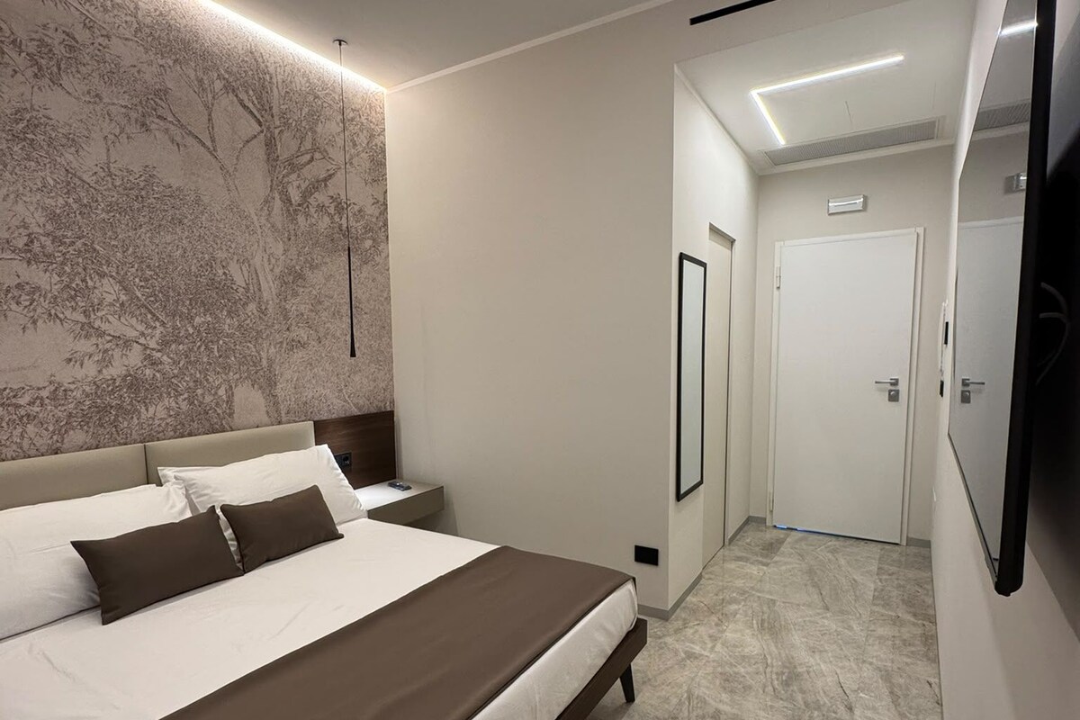 A modern bedroom with a large bed, a wall-mounted TV, and a hallway leading to a door.