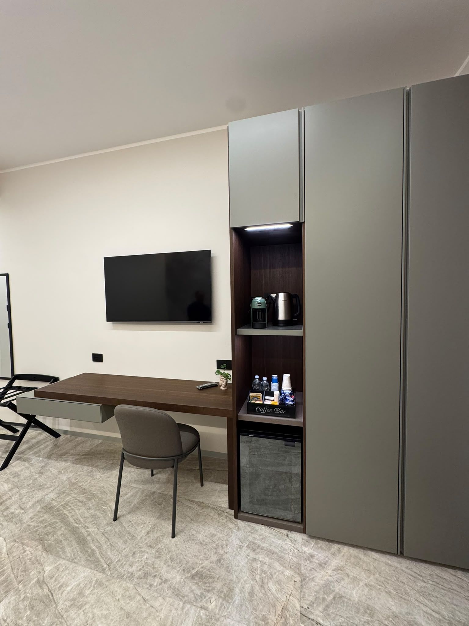 Un moderno spazio ufficio con una TV a schermo piatto montata a parete, una scrivania in legno con una sedia e un angolo caffè integrato con bollitore e macchina per il caffè.