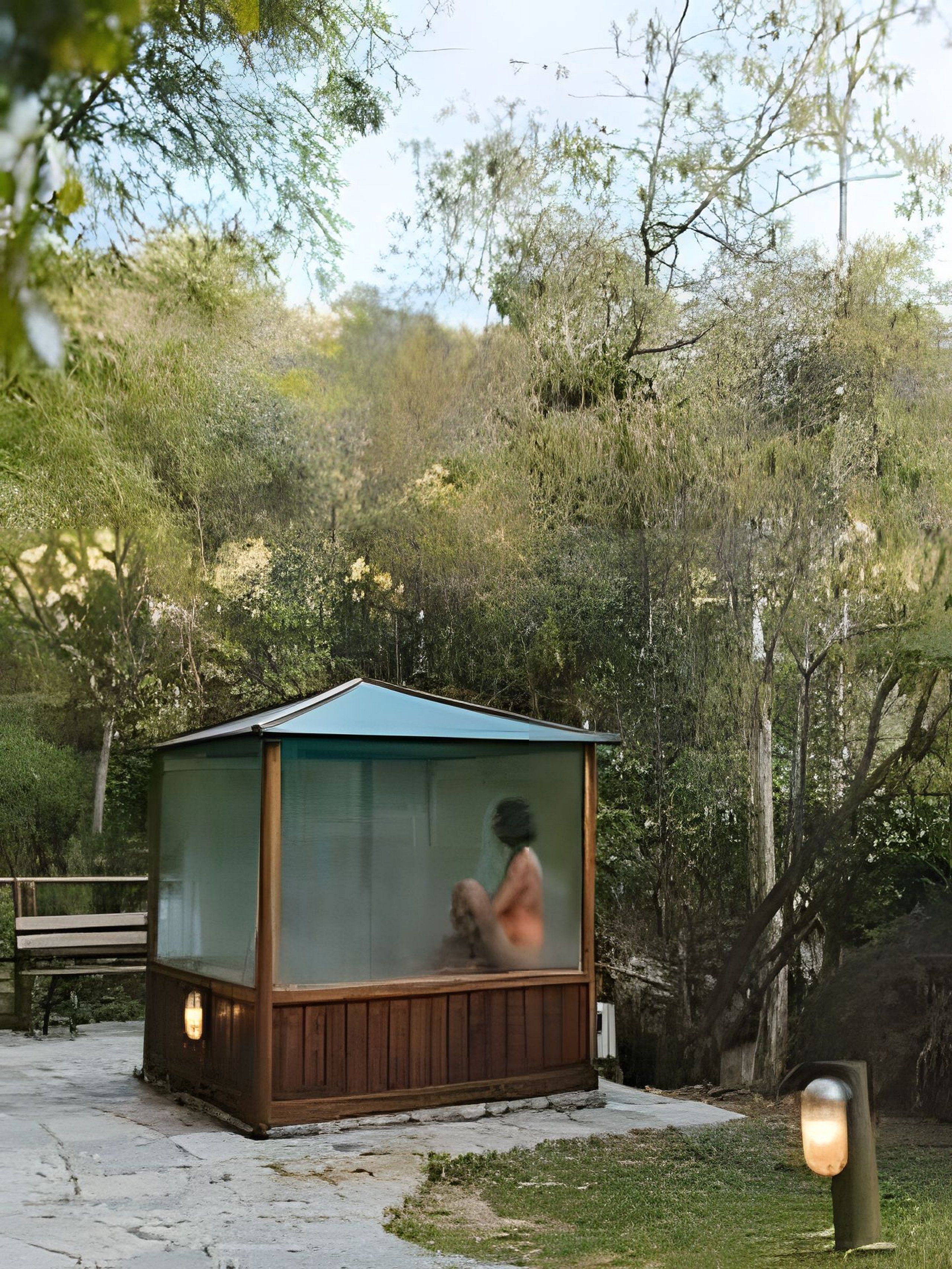 A small wooden structure with a glass enclosure, possibly a sauna or a small cabin, surrounded by trees and a paved area.