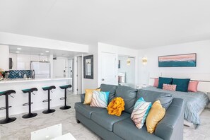 A modern living room with a grey sofa, a bar area with stools, a bed with a view, and a painting on the wall. - Renovated Beach View Condo, Fireworks, Waikiki (Honolulu)
