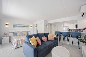 Un salon moderne avec un canapé, un coin repas doté de tabourets de bar, une cuisine et une chambre à coucher avec un lit et des tables de chevet.