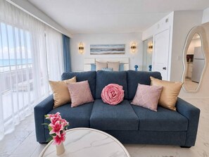 A modern living room with a blue sofa, decorative pillows, a vase with flowers, and a view of the ocean through large windows. - Lux Beach View Condo, Open 1BR, Waikiki (Honolulu)