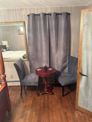 A room with a wooden floor, two grey upholstered chairs, a small round table with a red base, and a grey curtain.