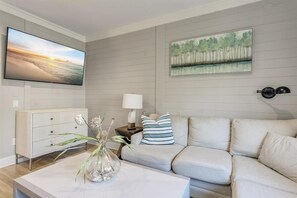 A living room with a flat-screen TV, a sofa, a coffee table with a vase, a painting on the wall, and a lamp on a side table. - Playa Maya (Panama City Beach)