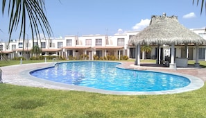 A swimming pool with a thatched-roof pavilion, surrounded by a well-maintained lawn and a row of buildings with a consistent architectural style.
