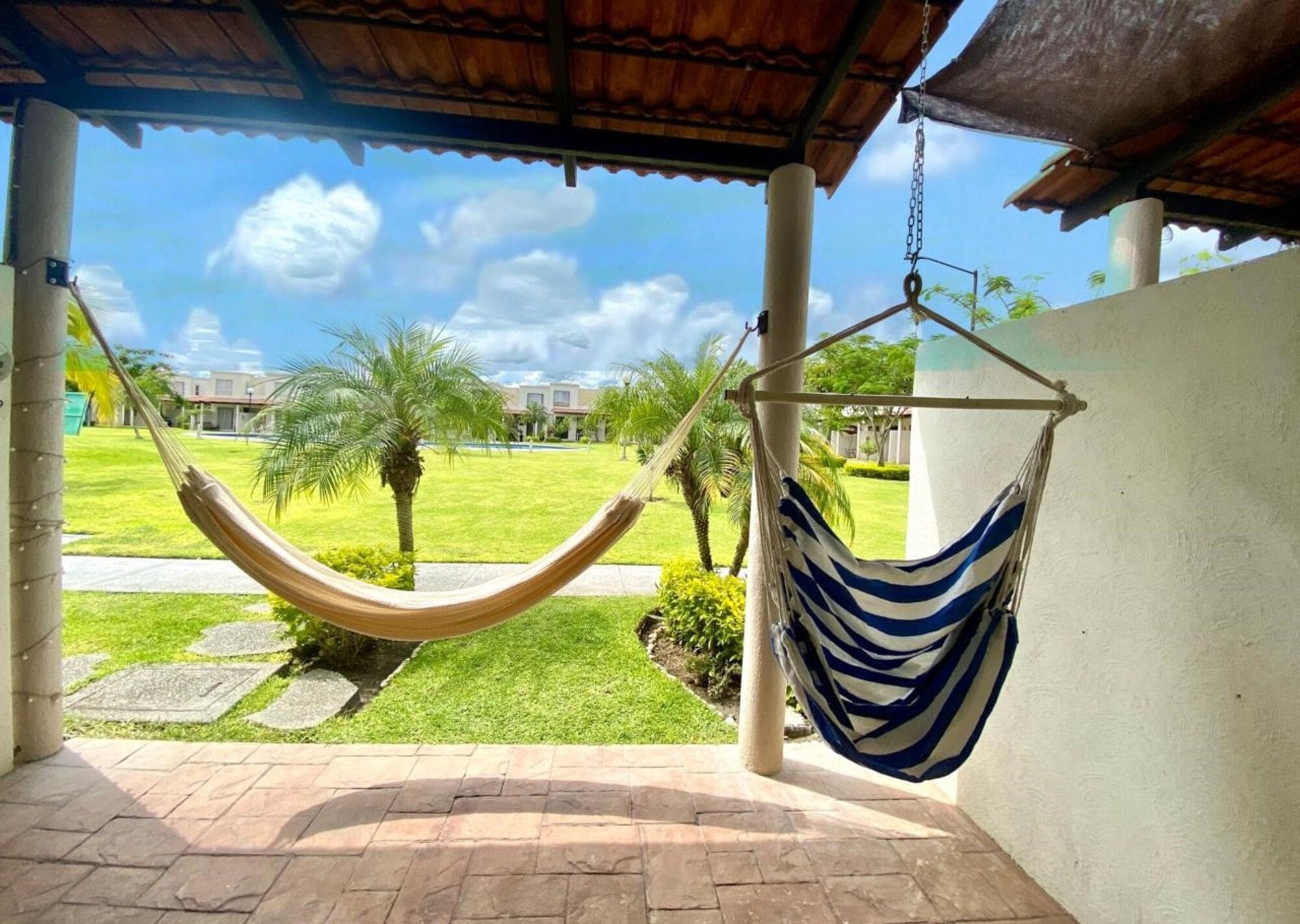 A hammock hanging between two columns in a covered outdoor area with a view of a well-maintained lawn and buildings in the background.