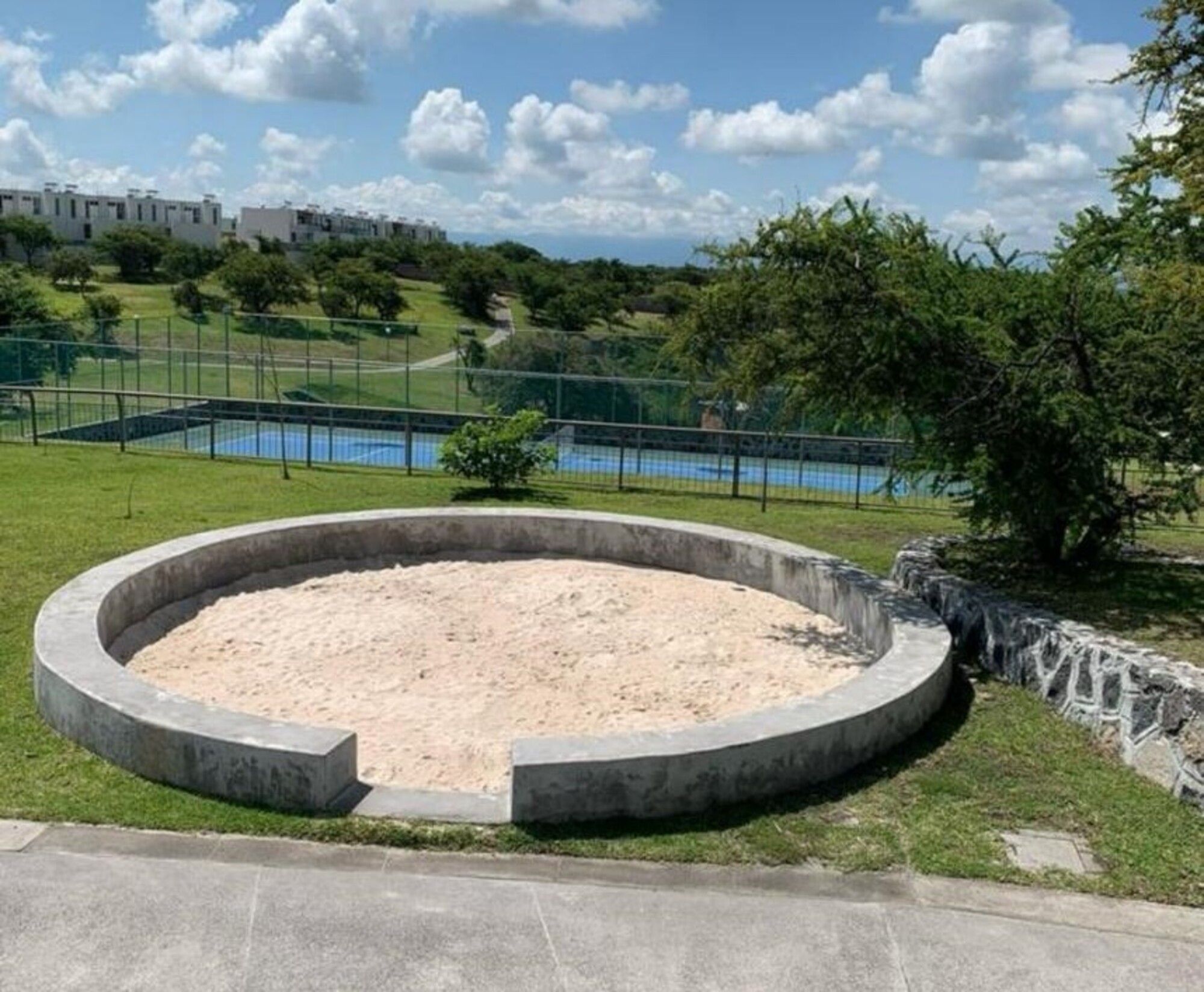 Eine runde Betonkonstruktion mit sandigem Boden, umgeben von einer Rasenfläche und einer Steinmauer.