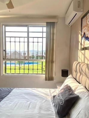 A hotel room with a bed, a window with a view of a pool and mountains, and an air conditioning unit.