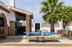 An outdoor patio with a ping pong table, chairs, a small table with a bowl of fruit, and a palm tree. - Villa Nerja | Pool | BBQ | Games | Fireplace (Nerja)