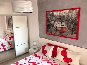 Habitación de hotel con cama, mesita de noche, espejo e imagen colgada en la pared de una bicicleta en un paisaje urbano.
