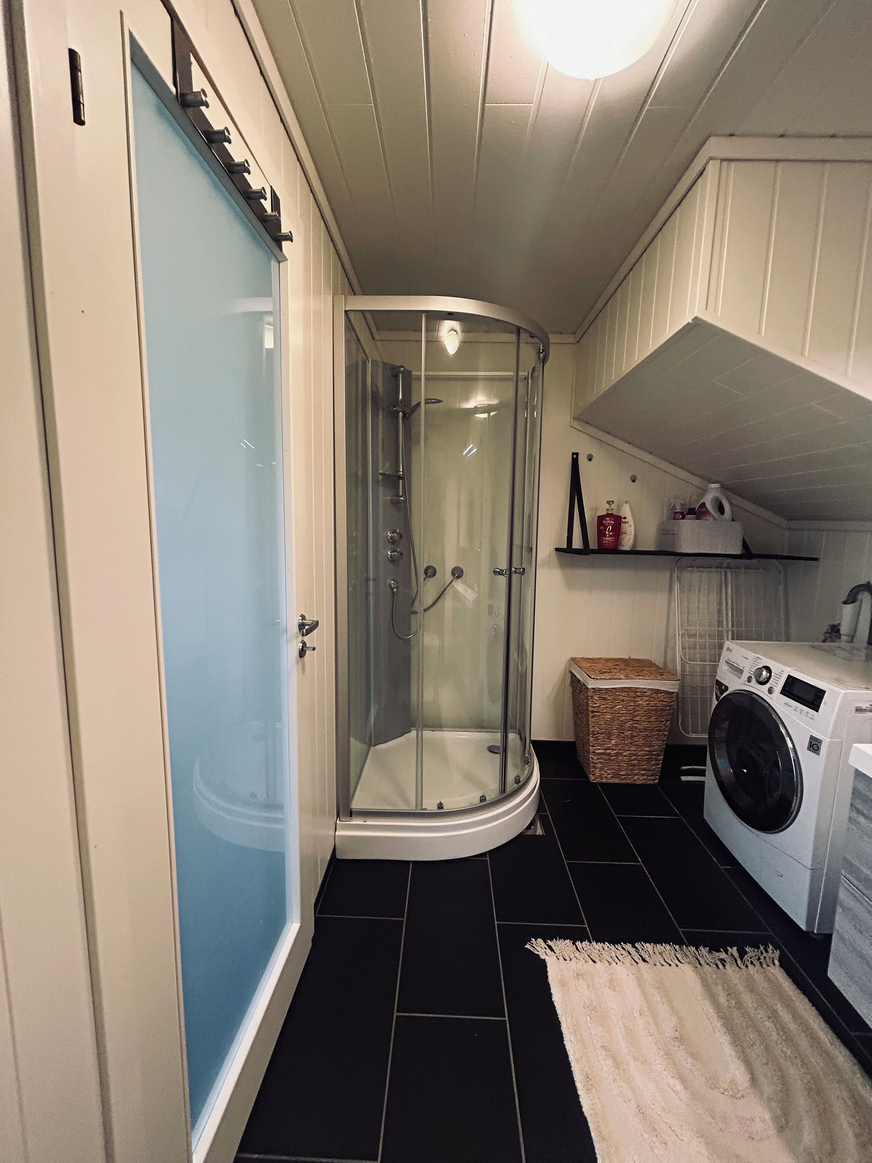 Een douchehoek met glazen deuren, een wasmachine en een wasmand in een kleine badkamer.