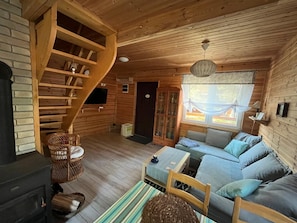 Ein gemütliches Blockhaus-Interieur mit einer Holztreppe, einer Sofagarnitur, einem Fernseher und einem Kamin.