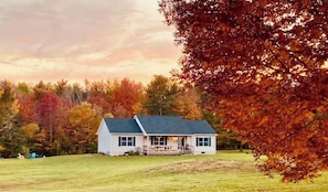A two-story house with a porch, surrounded by a grassy yard and trees with autumn foliage. - Coastal Farmhouse,10 min to beach, game room 1 dog (Lamoine)