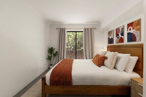 Ein Schlafzimmer mit einem hölzernen Bett, weißer Bettwäsche, einem Fenster mit Vorhängen und abstrakter Wandkunst.
