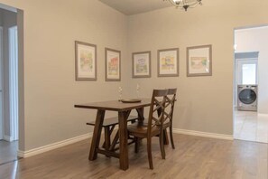 Un coin repas avec une table et des chaises en bois, des œuvres d’art encadrées au mur et un lave-linge à l’arrière-plan.