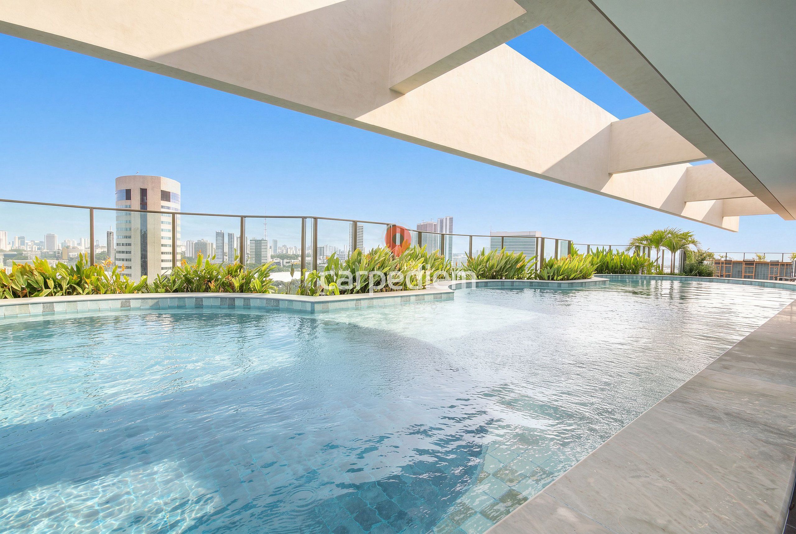 A rooftop swimming pool with clear blue water, surrounded by greenery and overlooking a cityscape.