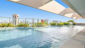 A rooftop swimming pool with clear blue water, surrounded by greenery and overlooking a cityscape.