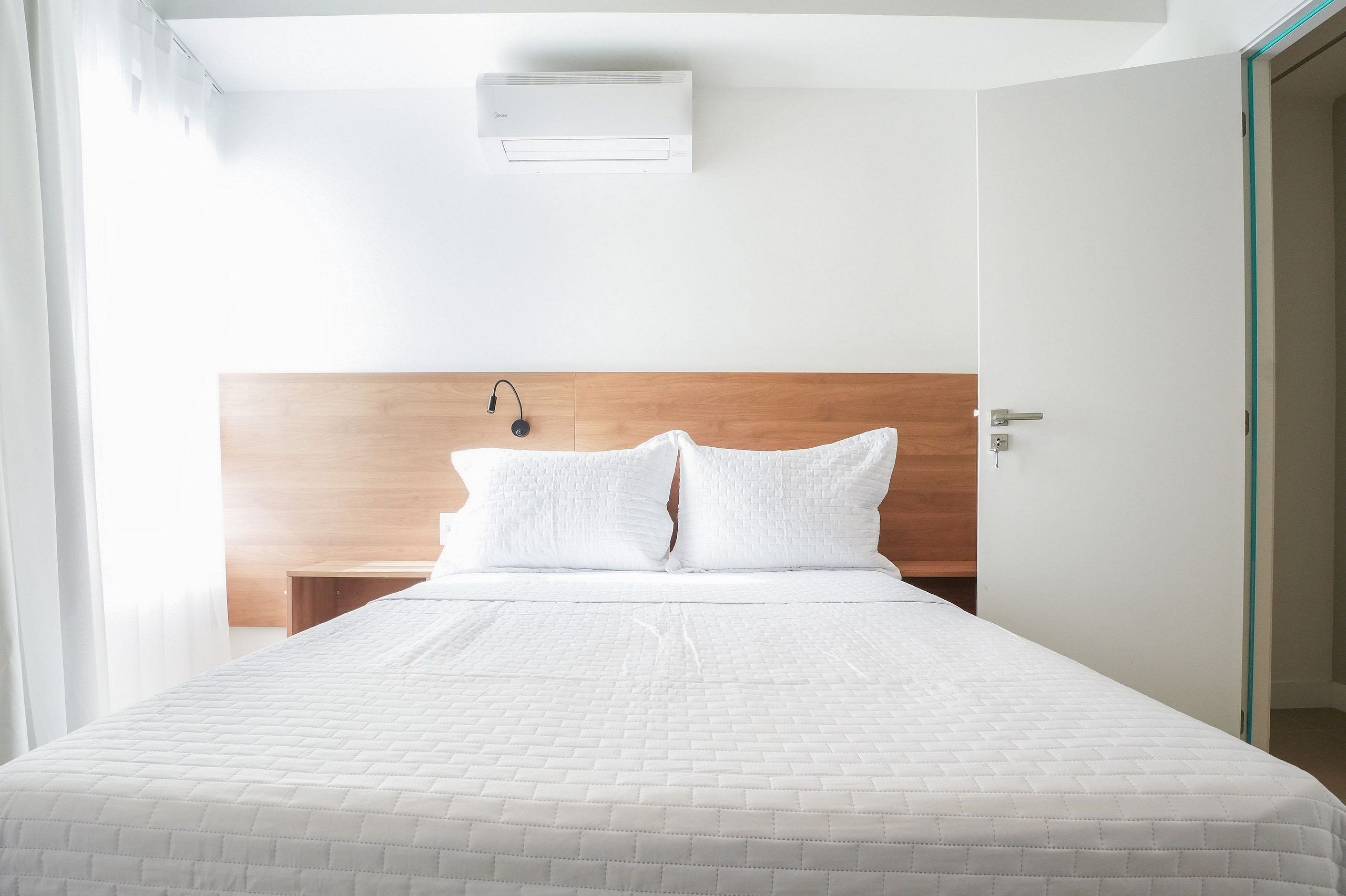 A hotel room with a bed, a wooden headboard, a wall-mounted air conditioner, and a door with a handle.