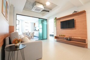 Un saló modern amb un sofà, una taula petita amb un ram, un televisor de pantalla plana muntat a una paret de fusta i una cuina visible a través d'una porta de vidre.