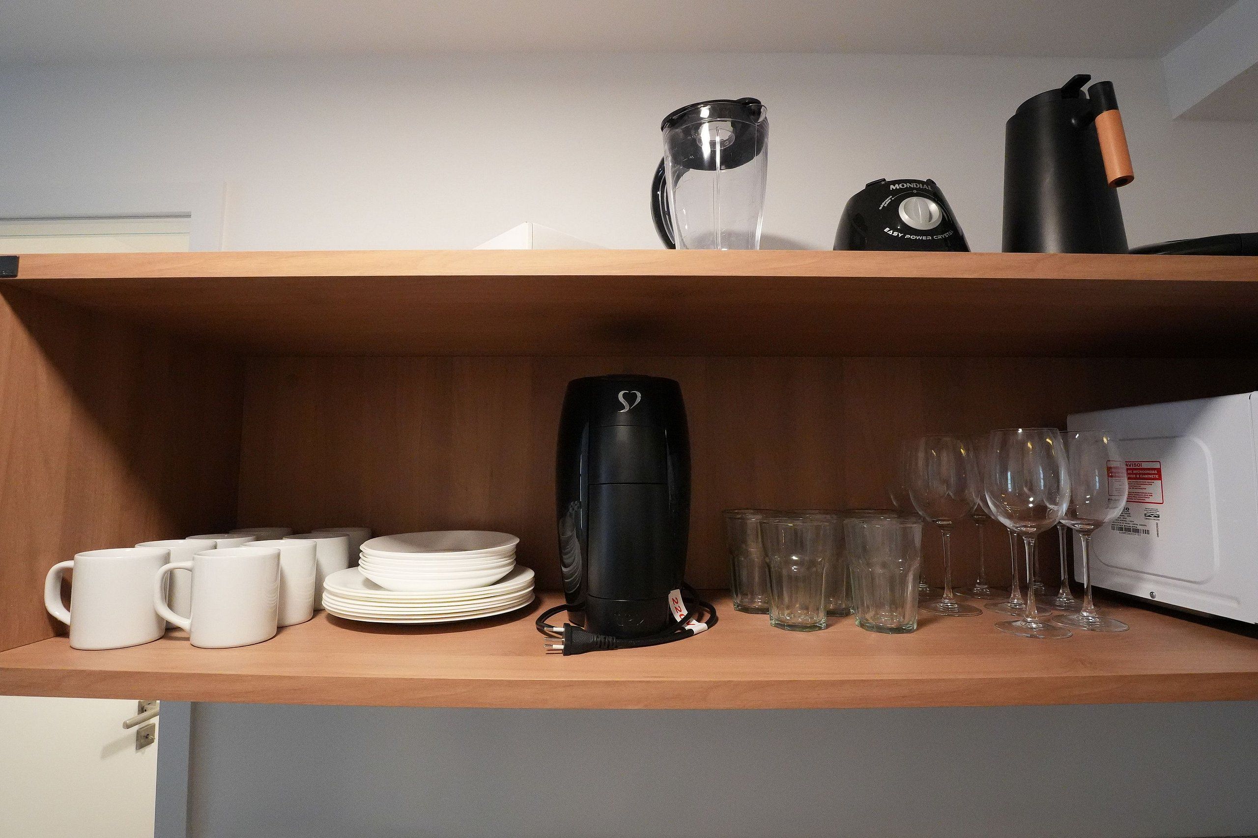 Un estante de cocina con tazas blancas, platos, una tetera eléctrica negra y copas de vino.