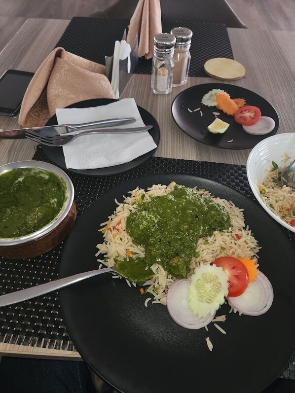 Una mesa servida con una comida que incluye arroz, verduras y una salsa verde, además de saleros, pimenteros, una servilleta y cubiertos.