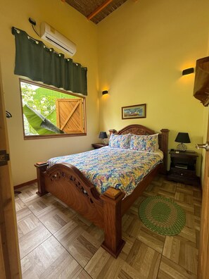 A bedroom with a wooden bed, a window with a view of greenery, a wall-mounted air conditioner, and a bedside table with a lamp. - Casa Bocaracá · Moderna · 4 PAX · Aire Acondicionado (Playa Chiquita)