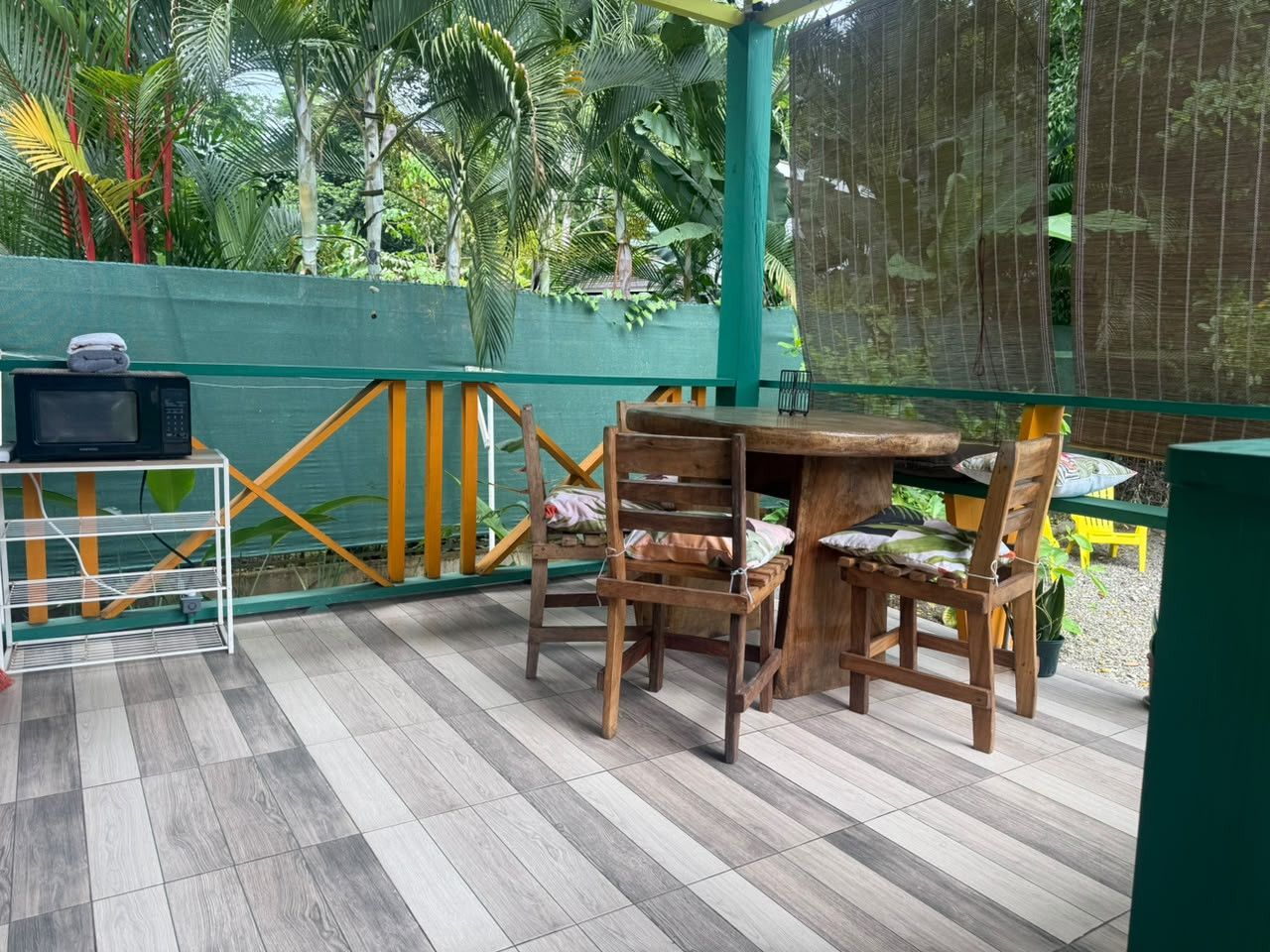 A wooden table and chairs set on a tiled patio with a green fence and tropical plants in the background.