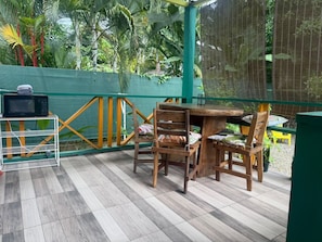 Een houten tafel met stoelen op een betegeld terras, met een groene hek en tropische planten op de achtergrond.