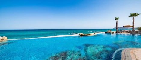 A clear blue pool with a view of the ocean, surrounded by rocks and palm trees.