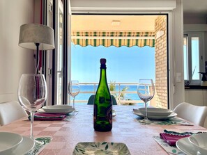 Ein Esstisch mit Flasche und Gläsern, Blick auf das Meer und ein Balkon mit gestreiftem Sonnensegel.