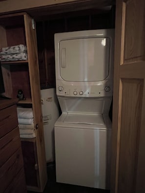 A compact, white, front-loading washing machine in a laundry room with wooden shelving and a small shelf.