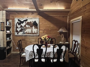 A wooden interior with a round table, chairs, a lamp, and a wall-mounted painting.