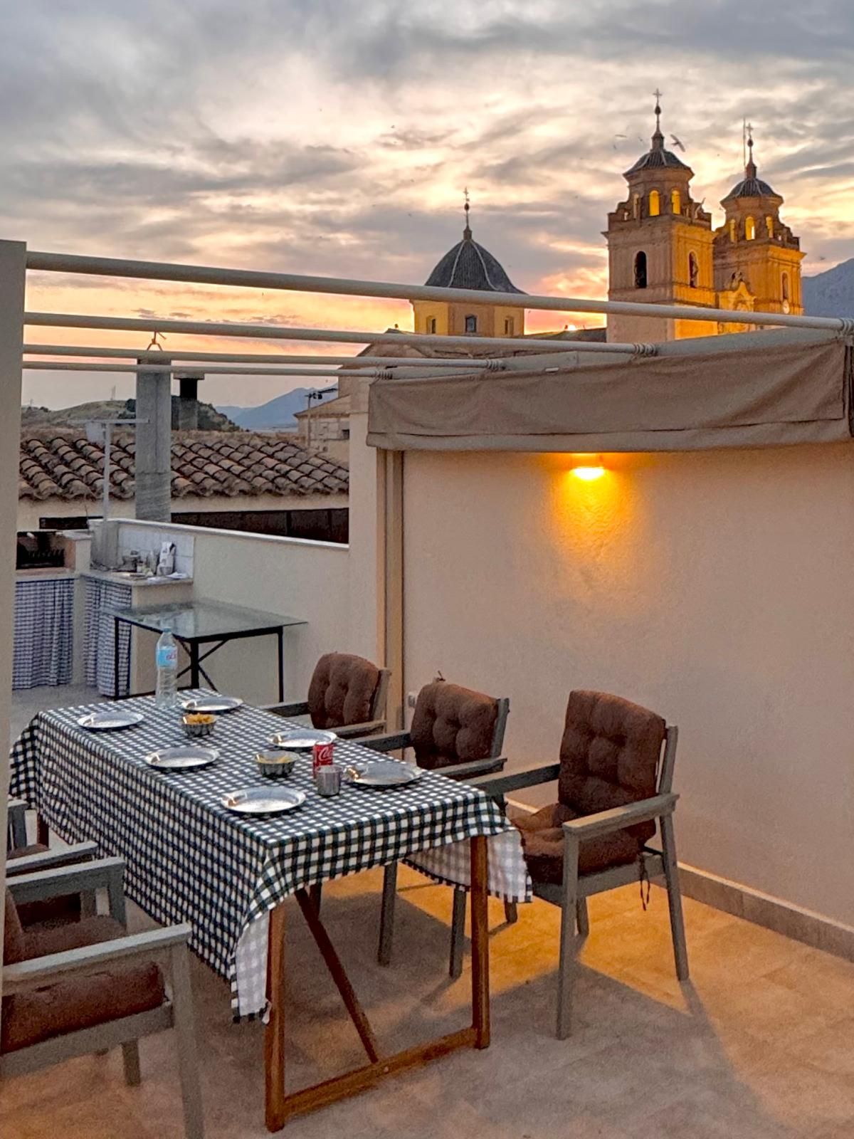 Un área de comedor al aire libre con una mesa dispuesta para dos personas, sillas y vistas a dos torres de iglesia al fondo.