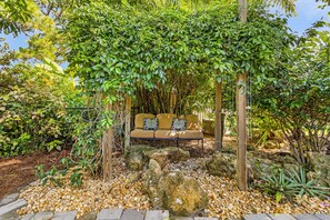 Eine hölzerne Bank mit Kissen unter einer mit Weinreben bewachsenen Pergola in einem Garten.