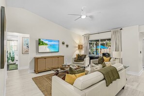 Ein Wohnzimmer mit einem weißen Sofa, einem Fernseher, einem Deckenventilator und Blick ins Freie durch eine Glastür.