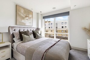 Ein Schlafzimmer mit einem großen Bett, einem Nachttisch, einer Kommode, einem Fenster mit Blick auf Gebäude und einer Deckenleuchte.