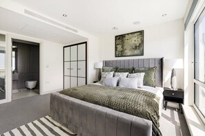 Ein modernes Schlafzimmer mit einem großen Bett, einer grauen Kopfteil und Blick auf die Stadt durch ein großes Fenster.