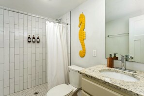 Un baño con ducha de azulejos blancos, un inodoro, un lavabo de encimera de granito y un espejo.