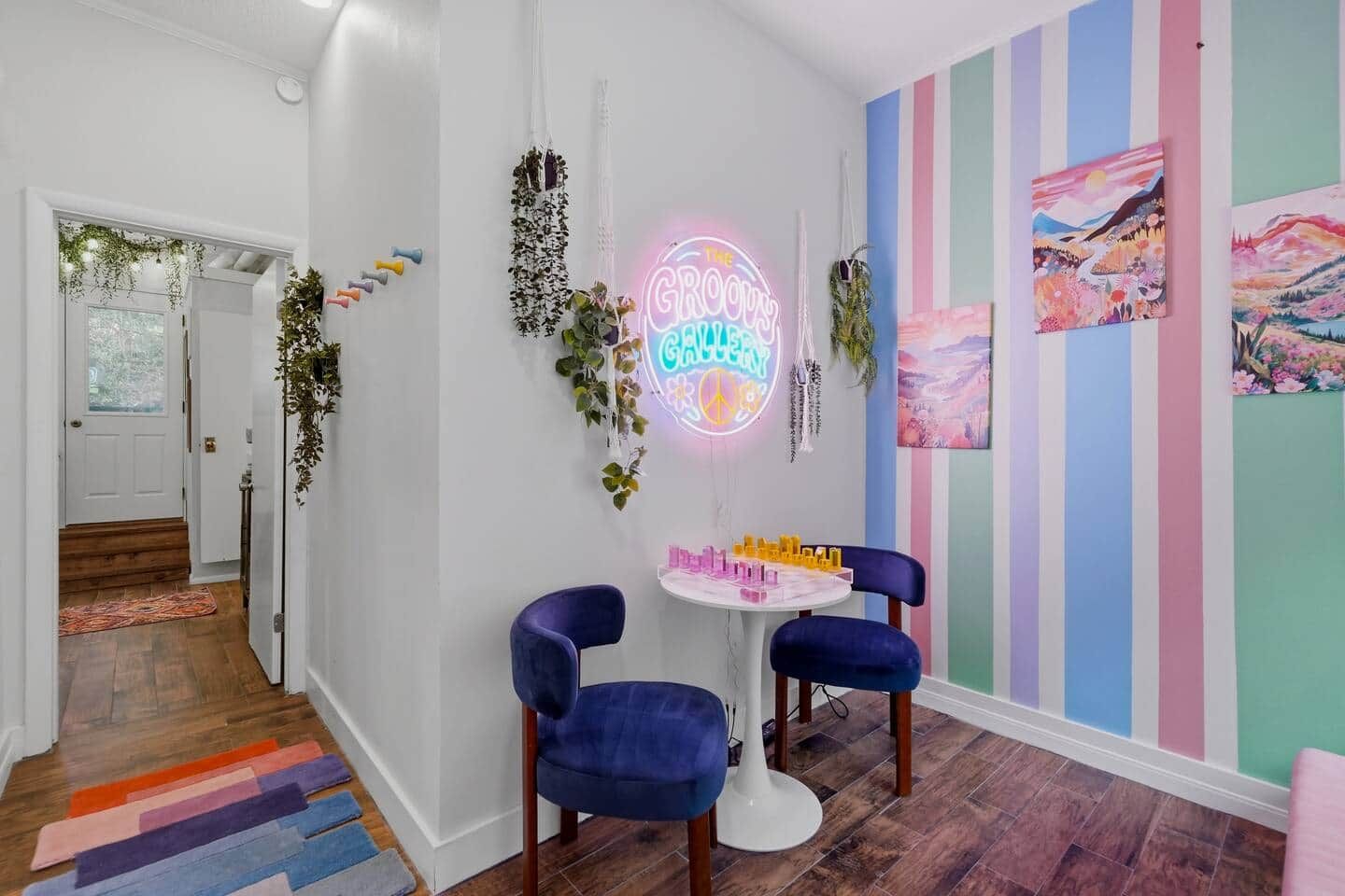 A hallway with a neon sign, two blue chairs, a small table with bottles, and a colorful striped wall.