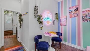 A hallway with a neon sign, two blue chairs, a small table with bottles, and a colorful striped wall.