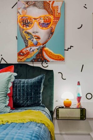 Un dormitorio con cama, almohadas, una mesita de noche con lámpara y una obra de arte en la pared.
