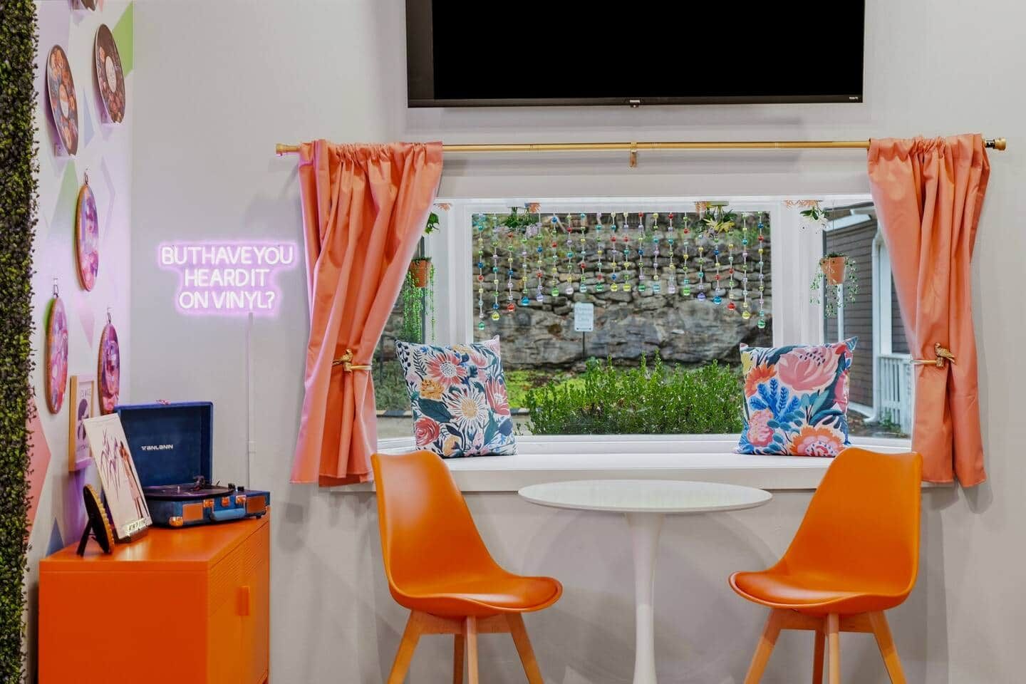 Een kamer met een aan de muur bevestigde televisie, een raam met gordijnen, twee oranje stoelen, een klein tafeltje en een neonbord.