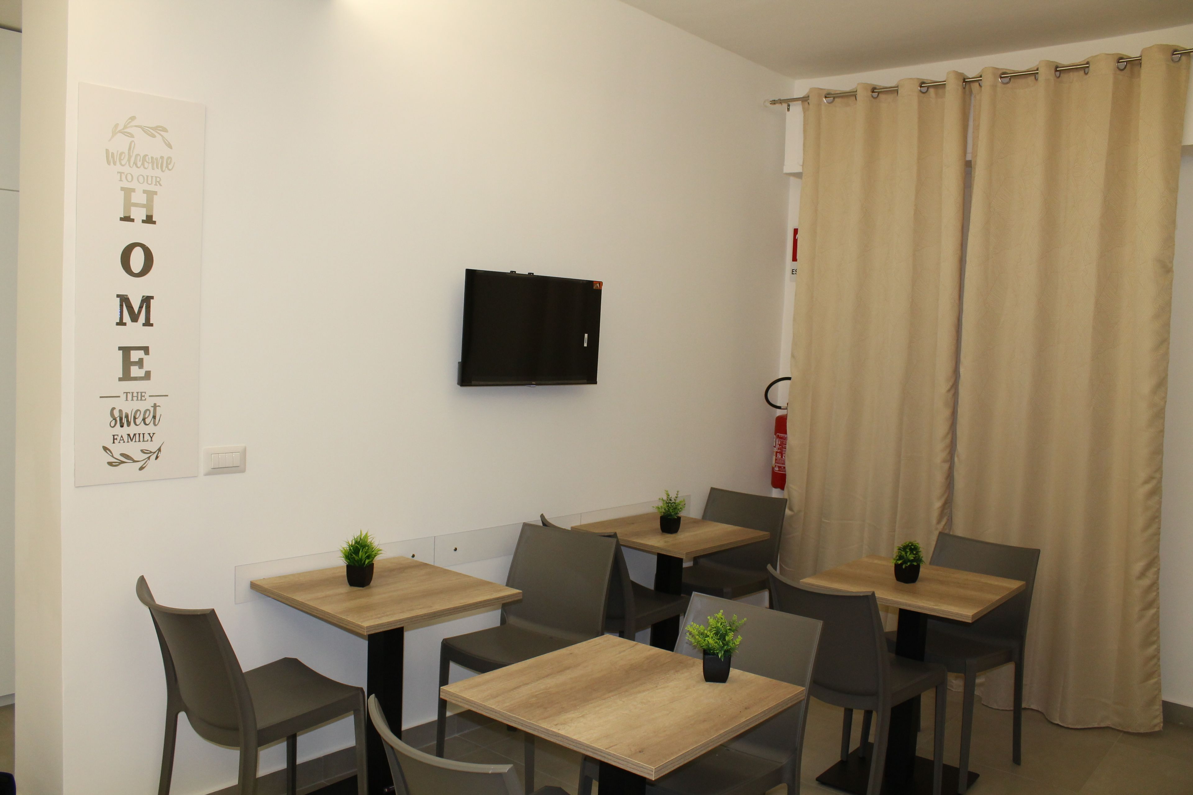Una habitación pequeña y moderna con una televisión de pantalla plana en la pared, una pizarra con un mensaje de bienvenida y una ventana con cortinas color beige.