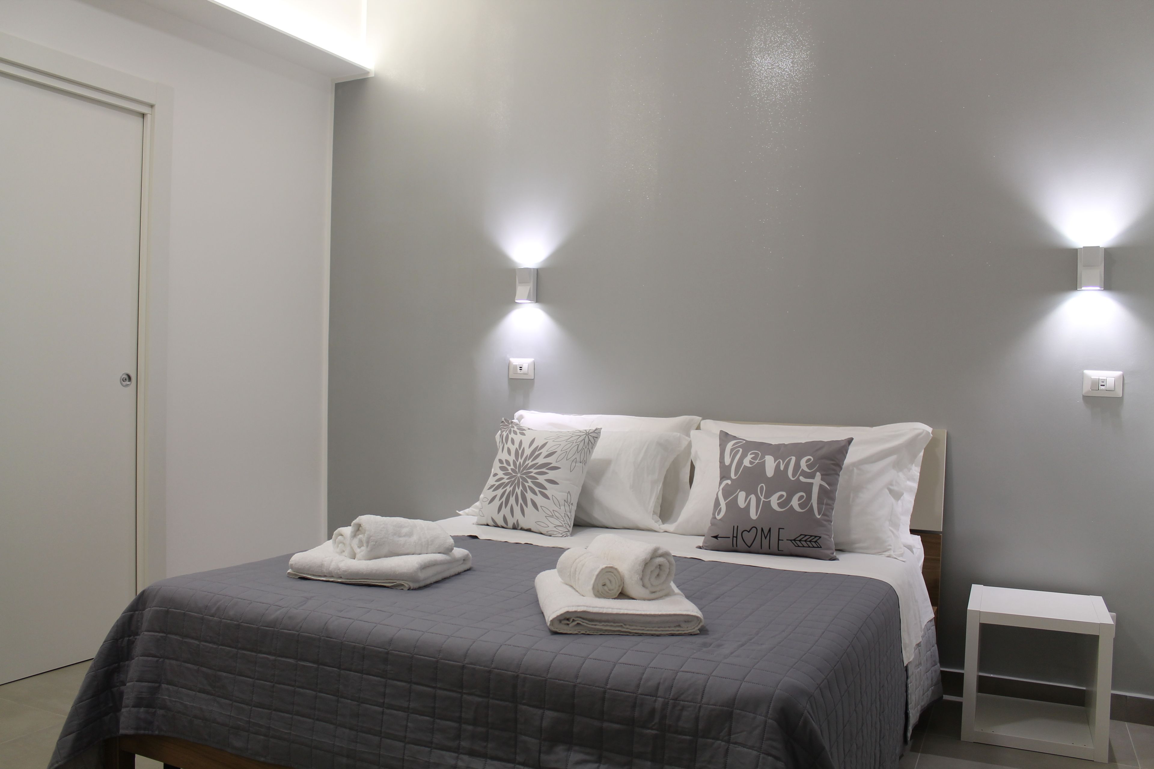 A bedroom with a bed, two wall-mounted lights, a bedside table, and a door.