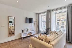 Ein modernes Wohnzimmer mit einer Couch, einem Fernseher, einer weißen Ablage und einem großen Fenster mit Vorhängen.