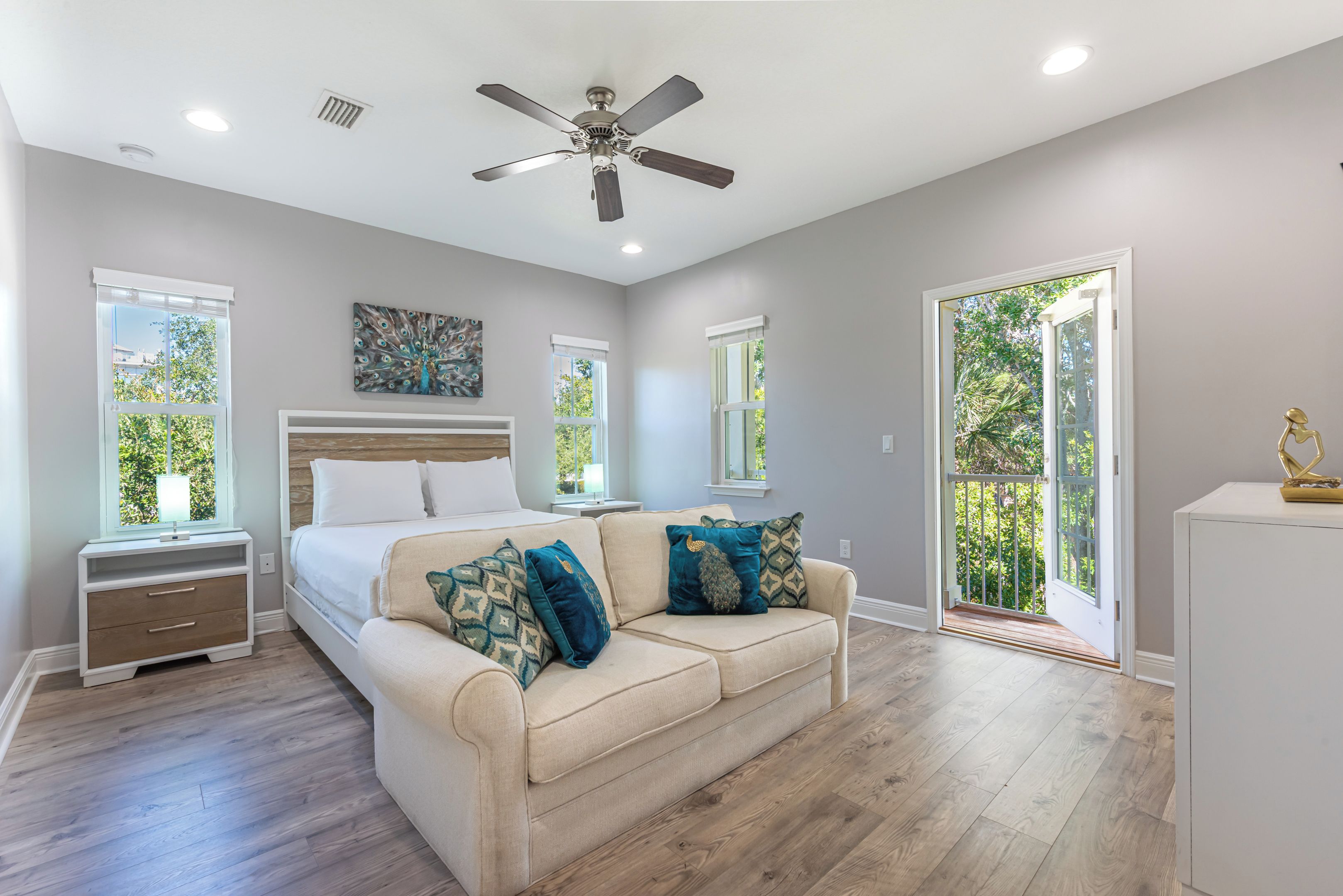 A bedroom with a bed, a sofa, a ceiling fan, and a window with a view of greenery.